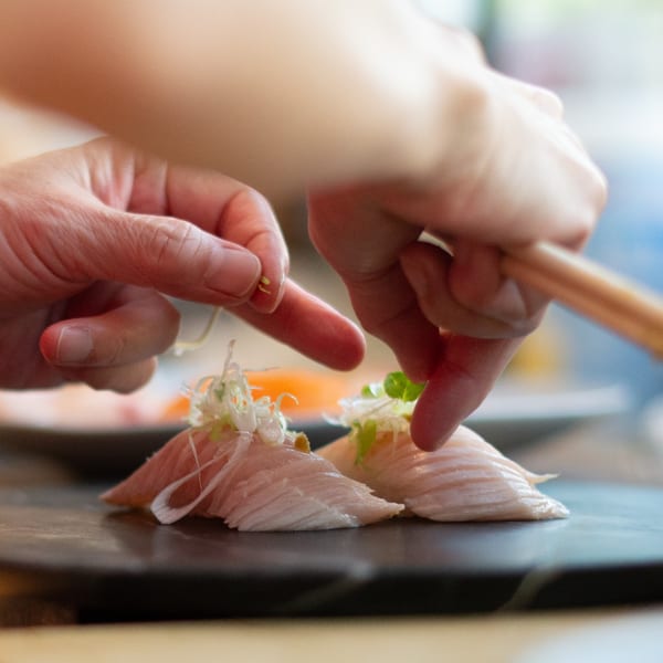 Chef's hands meticulously garnishing two pieces of delicate Pisces sushi with fresh herbs on a dark plate.