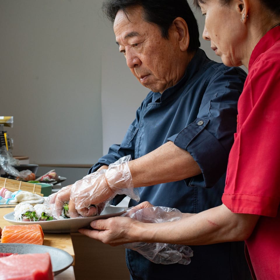 Two chefs, one in blue and one in red, preparing sushi rolls on a white plate.