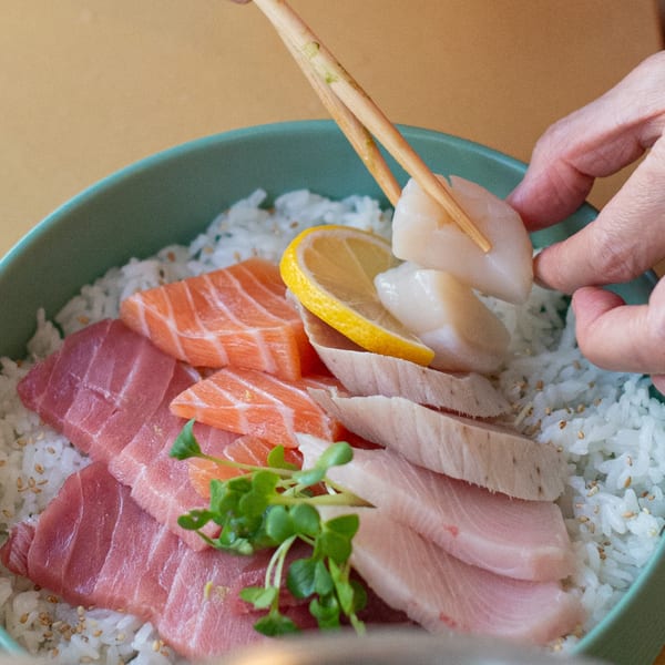 A hand places a scallop with chopsticks onto a Pisces sushi bowl with salmon, tuna, and lemon.