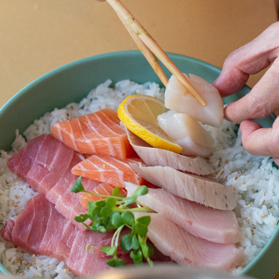 A hand places a scallop with chopsticks onto a Pisces sushi bowl with salmon, tuna, and lemon.