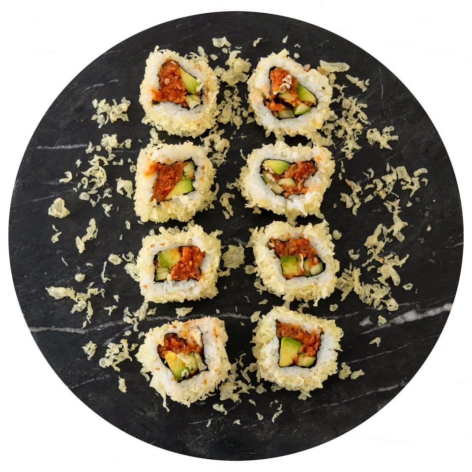 Eight Pisces sushi rolls with red filling and avocado, sprinkled with tempura flakes on a dark plate.