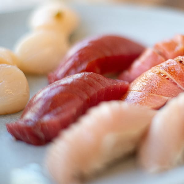 Close-up of various sushi pieces, including red tuna and lighter-colored scallops, on a white plate.