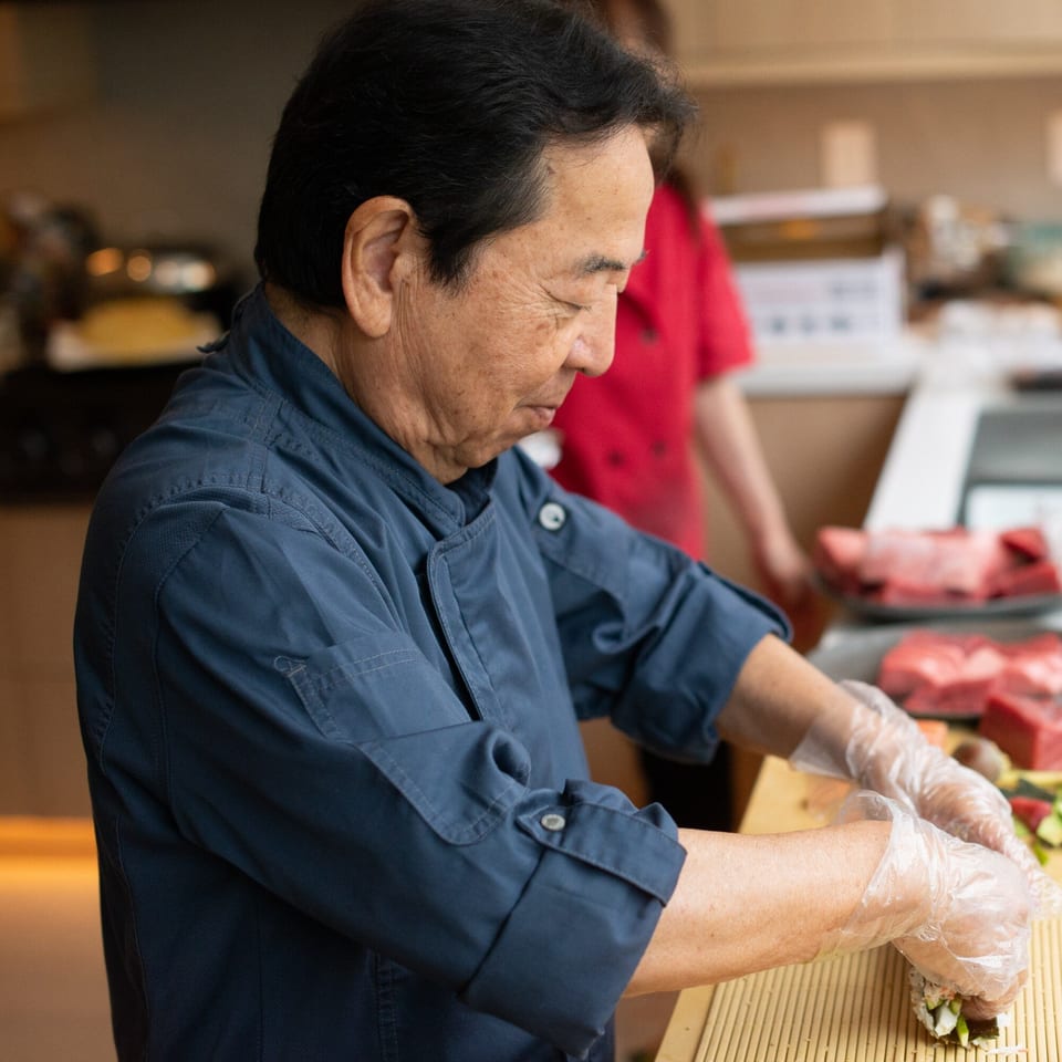 A Pisces sushi chef in a blue uniform and gloves carefully rolls a sushi creation.