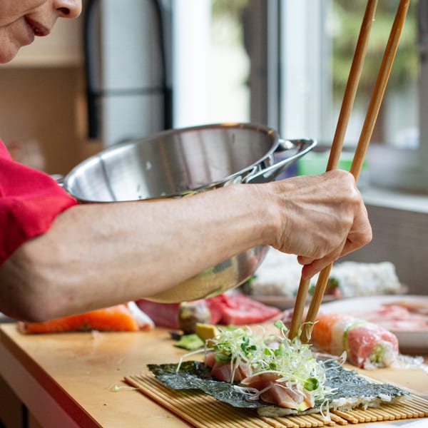 A chef uses chopsticks to add garnishes to a sushi roll on a bamboo mat.