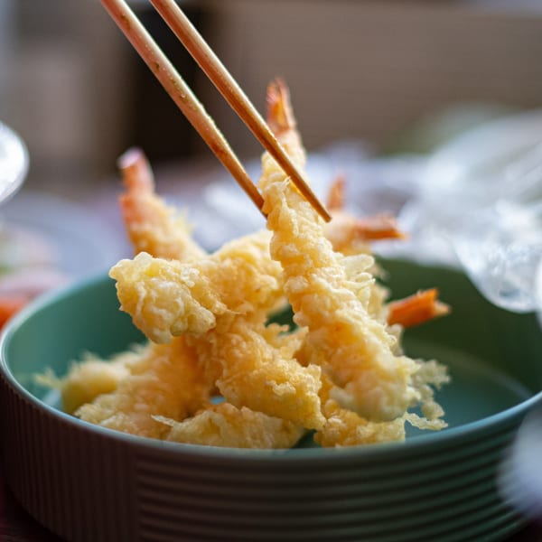 Chopsticks picking up a piece of crispy shrimp tempura from a teal bowl at Pisces.